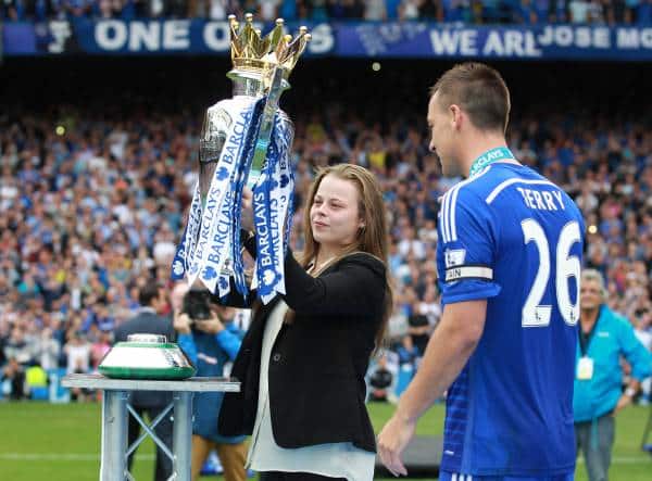 Young woman triumphantly holds Premier League trophy, surrounded by celebrating fans.
